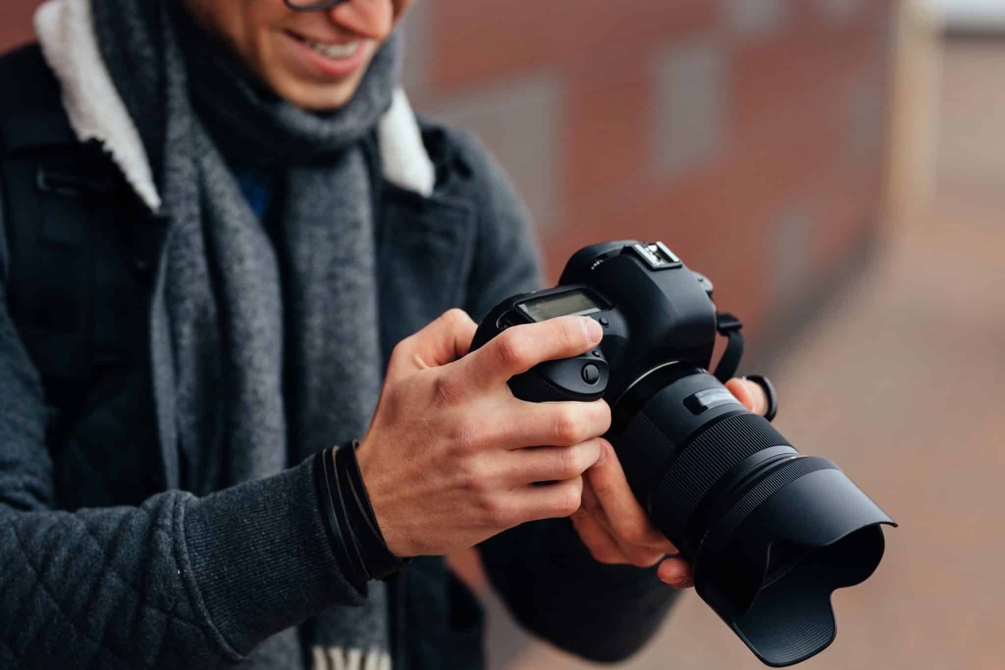 Ein fröhlicher junger Mann betrachtet Fotos in der Kamera. Gekleidet in eine warme, stilvolle Jacke und einen grauen Schal. Steht auf der Straße.