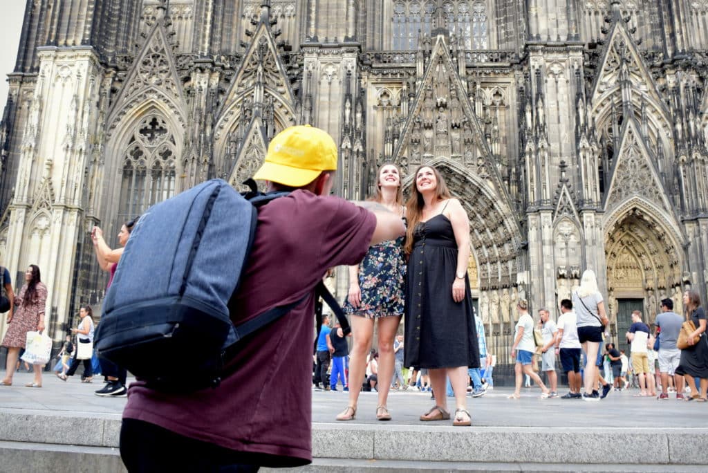 Dieses Foto zeigt einen Fotografen mit einer auffälligen gelben LET IT CLICK Cappi, der zwei Freundinnen vor dem beeindruckenden Kölner Dom an einem schönen Tag fotografiert. Der Dom