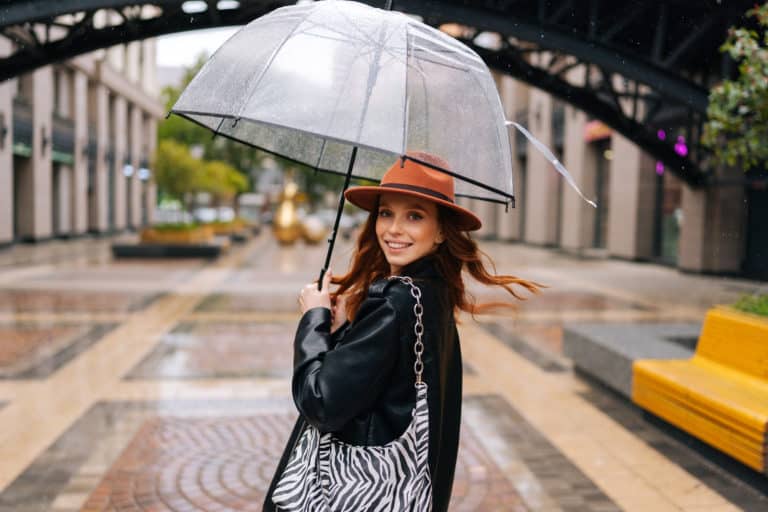 Porträtfotos: Elegante Frau mit modischem Hut und transparentem Regenschirm auf einer Stadtstraße, ein Symbol für Fotografie und weiblichen Lifestyle.
