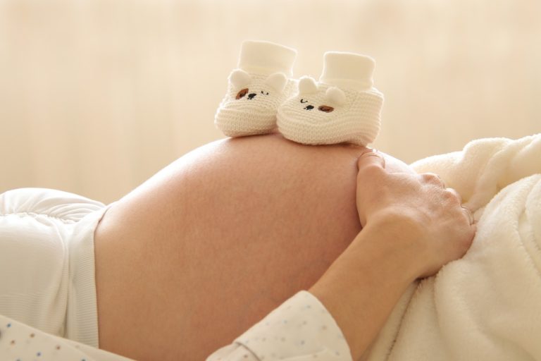 Eine werdende Mutter liegt entspannt auf dem Rücken, während der Fokus auf ihrem schönen Babybauch liegt. Auf dem Bauch liegen zwei kreative Babyschuhe mit charmanten Tiergesichtern, die die einzigartig schöne Vorfreude auf das neue Familienmitglied perfekt einfangen.