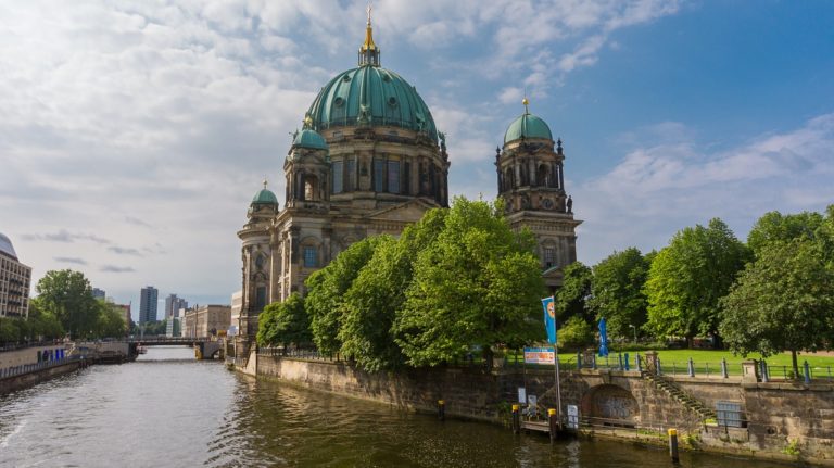 Der Berliner Dom, von der Spree aus betrachtet. Auf der rechten Seite des Bildes säumen einige Bäume das Ufer, während die imposante Kuppel des Doms im Zentrum steht und sich im ruhigen Wasser der Spree spiegelt.