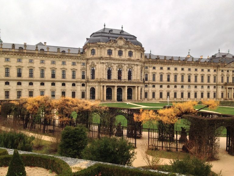 Ein herbstlicher Blick auf die Würzburger Residenz, eingefasst von einer kleinen Allee mit goldenen Blättern, die den majestätischen Barockbau sanft umrahmen.