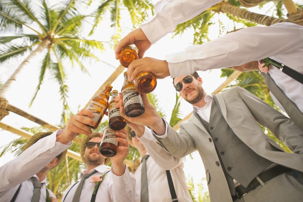 Mehrere Männer in eleganten Anzügen stoßen mit Bierflaschen an, während sie unter Palmen im Freien feiern. Die entspannte Atmosphäre und das sonnige Wetter sorgen für einen lässigen Junggesellenabschied oder eine Feier im tropischen Stil.