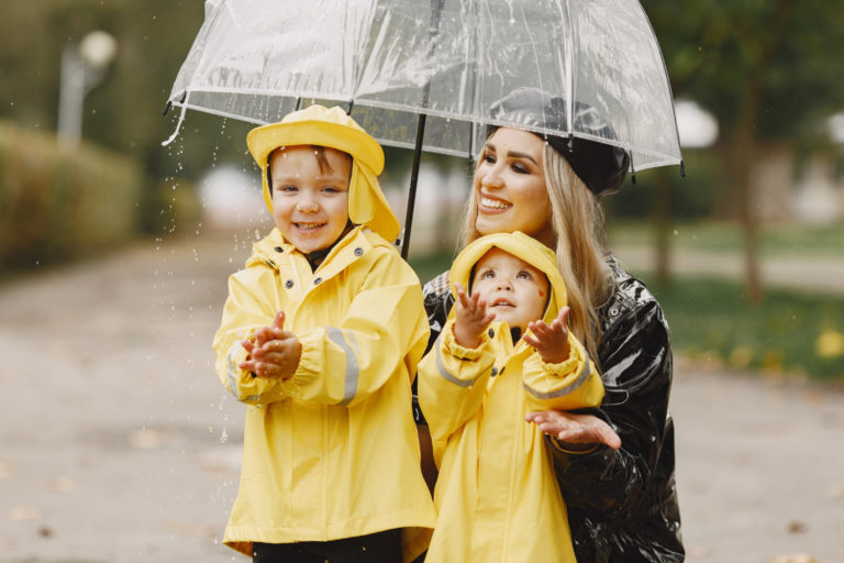 Mutter und zwei Kinder in gelben Regenjacken genießen ein Fotoshooting im Regen unter einem transparenten Regenschirm, fangen lachend den Regen.