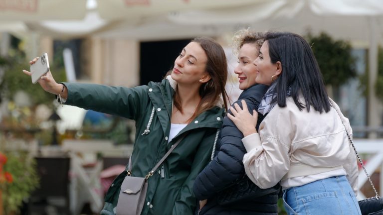 Drei junge Frauen posieren für ein Selfie im Freien, eine von ihnen hält ein Smartphone, um das Foto aufzunehmen. Sie lachen und stehen eng beisammen.