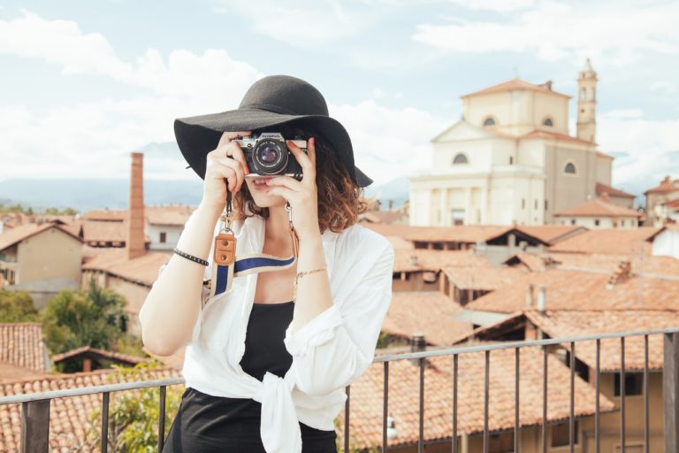 Frau mit schwarzem Hut und weißer Bluse hält eine Kamera vor ihr Gesicht, während sie in einer Stadt mit rustikalen Gebäuden und blauen Himmel fotografiert.