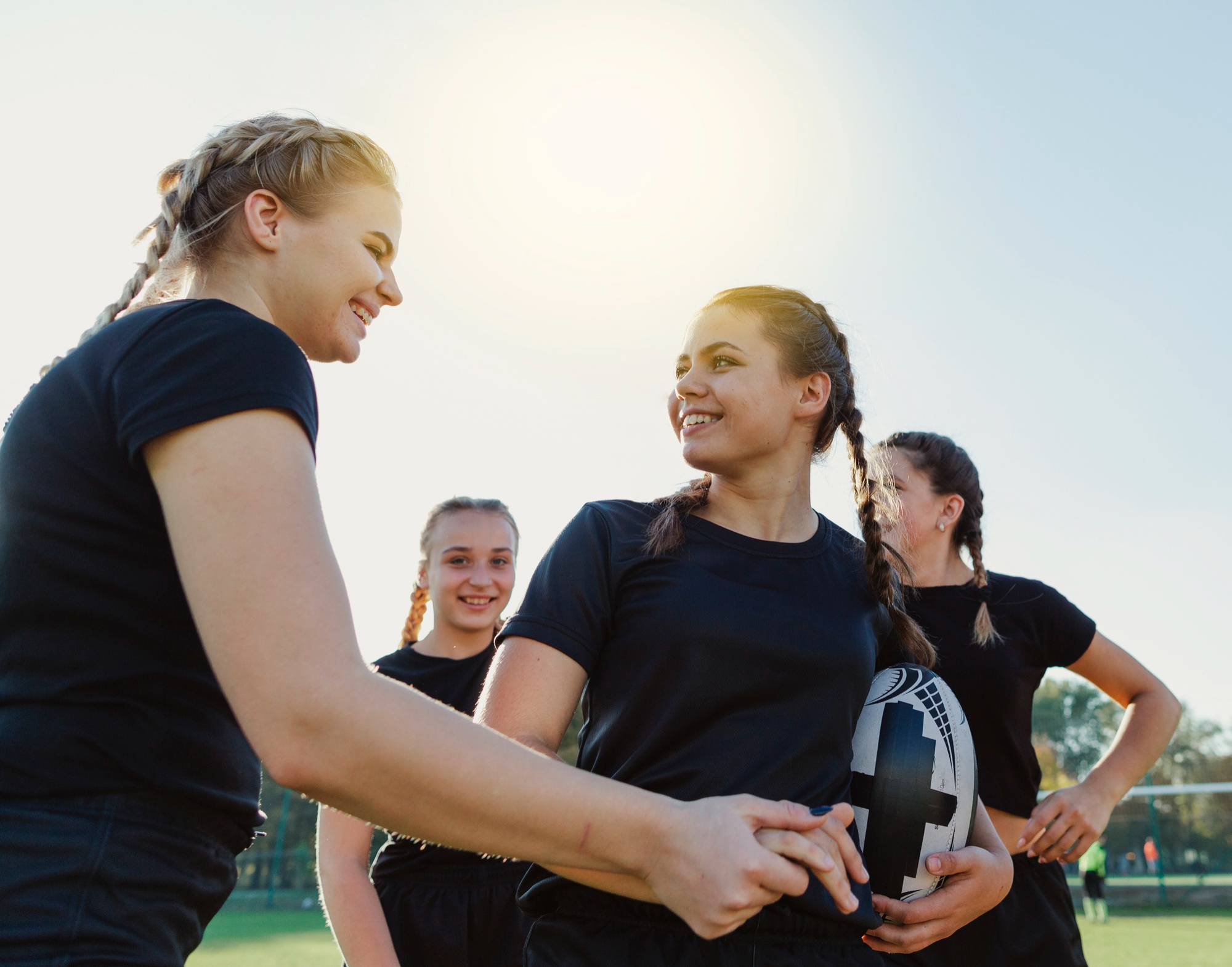 Vereinsfotos voller Teamgeist: Eine Gruppe junger Frauen im Sportoutfit lacht gemeinsam auf dem Spielfeld, während eine von ihnen einen Rugbyball hält.