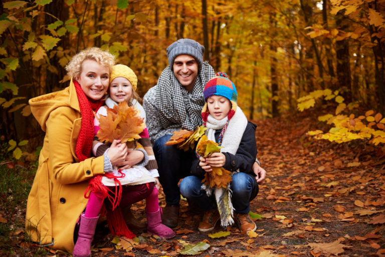 Familie mit zwei Kindern beim Outdoor-Shooting im herbstlichen Wald, umgeben von bunten Blättern und in warmen Herbstkleidern lachend zusammen.