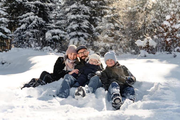 Familie sitzt eng beisammen im Schnee und lächelt in winterlicher Waldkulisse für Familienfotos im Schnee.