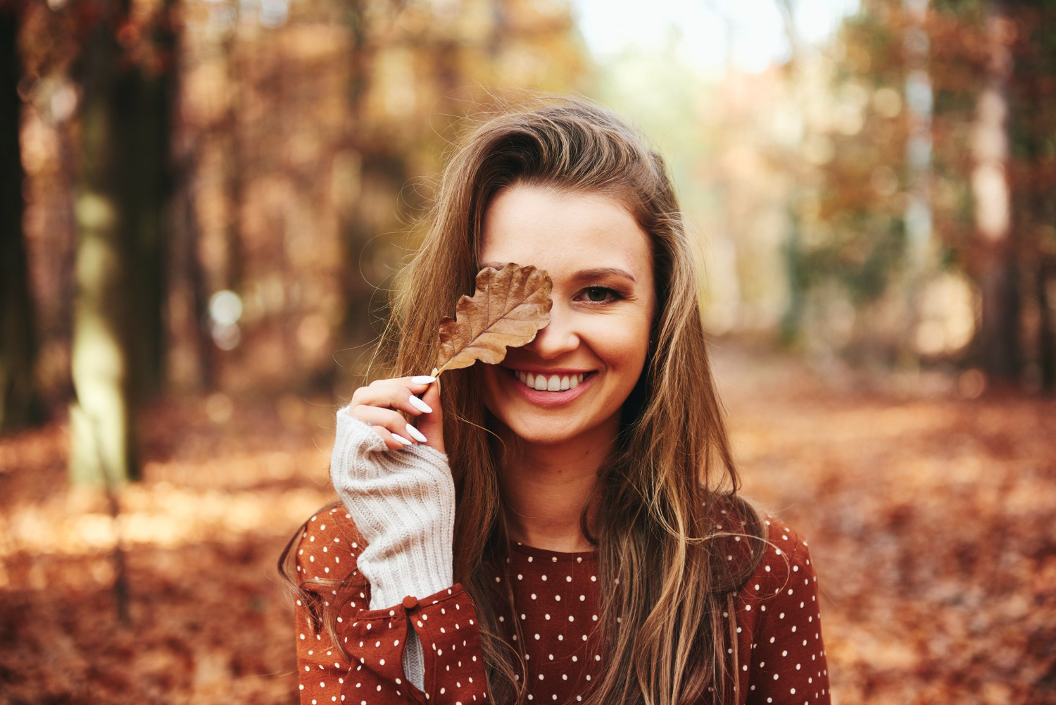 Junge Frau lächelt bei einem Outdoor-Shooting im Herbst, hält ein Eichenblatt vor ihr Gesicht und steht in einem herbstlichen Wald.