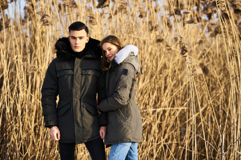 Paar in warmen Jacken posiert bei einem Outdoor-Shooting im Herbst vor hohem, trockenem Gras und goldener Naturkulisse.