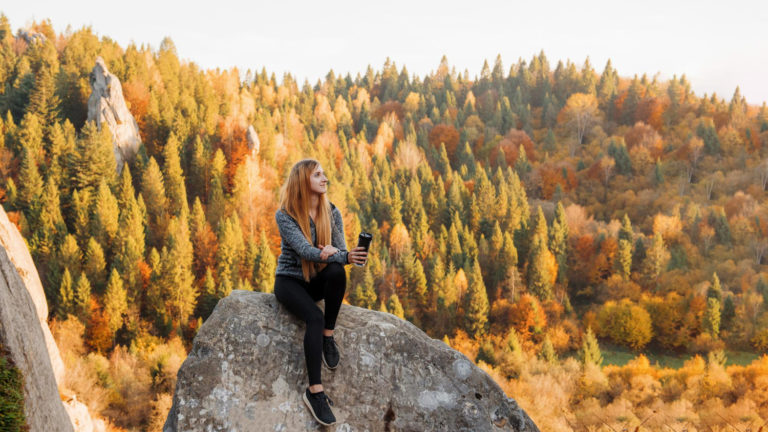 Frau sitzt auf einem Felsen bei einem Outdoor-Shooting im Herbst, umgeben von farbenfrohem Wald und Berghängen in herbstlichen Farben.