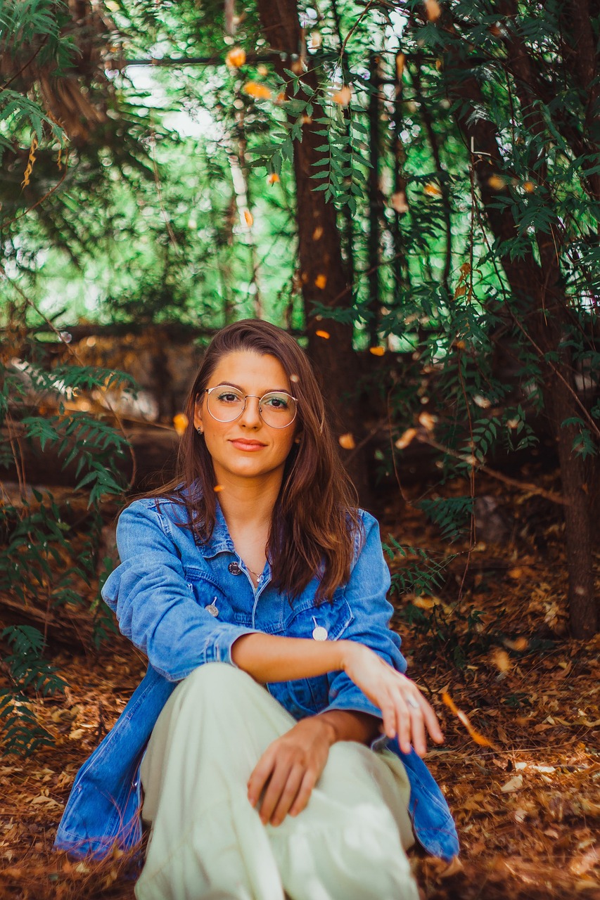 Junge Frau in Jeansjacke sitzt im herbstlichen Wald, umgeben von fallendem Laub, entspannte und natürliche Atmosphäre.