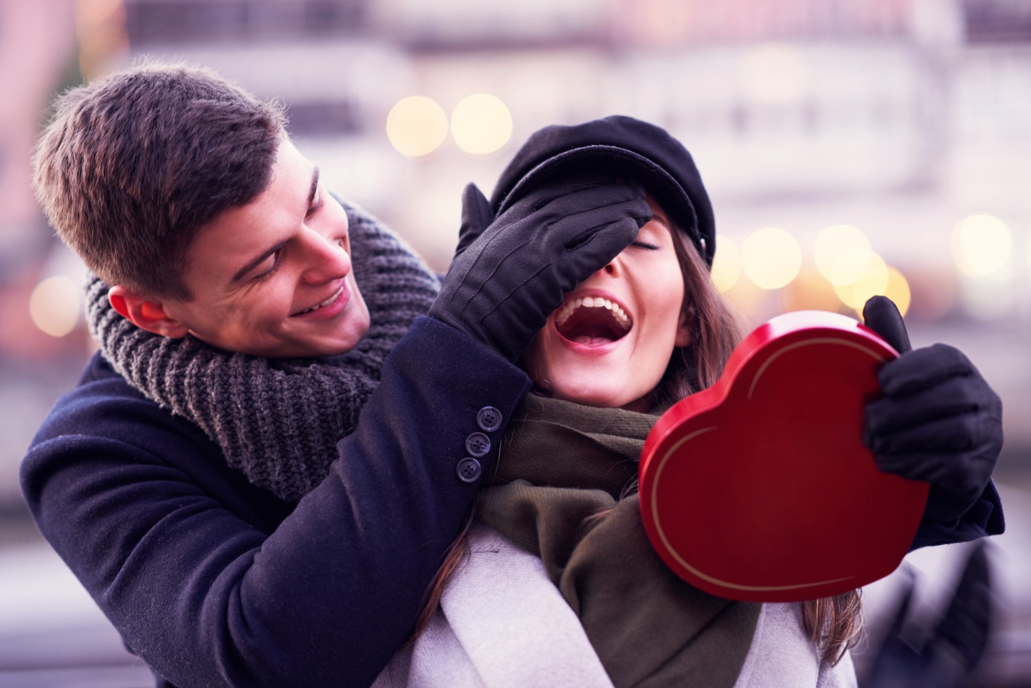 Ein lachendes Paar während eines Valentinstag-Fotoshootings – der Mann überrascht die Frau mit einer roten Herzbox.