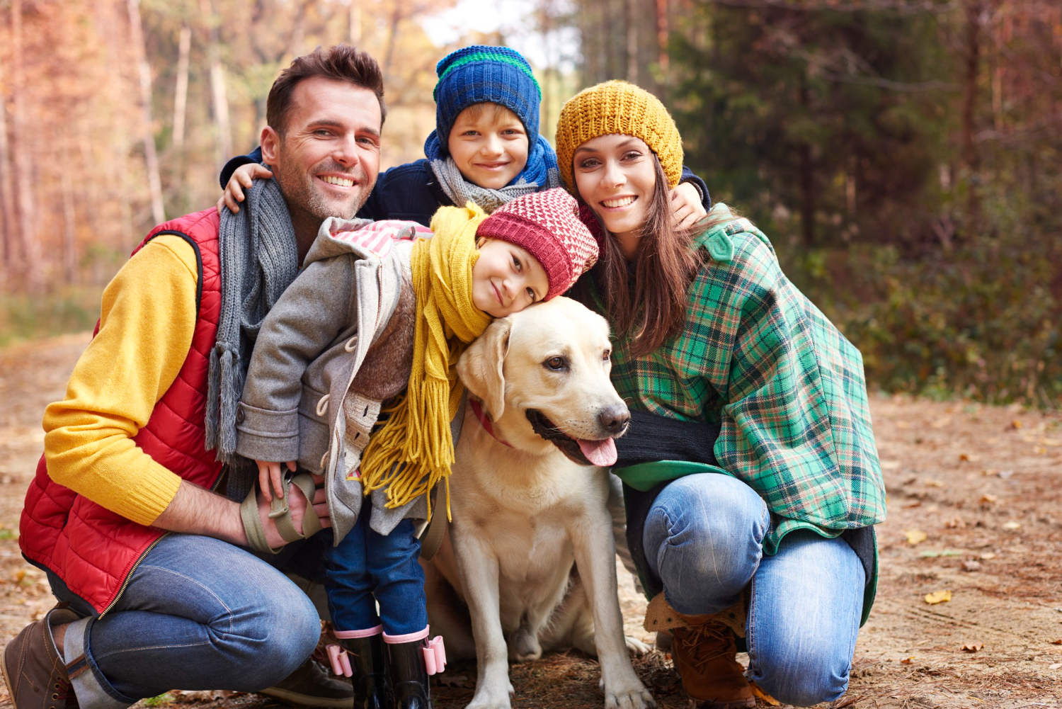 Eine lächelnde Familie posiert zusammen mit ihrem Hund während eines Herbstspaziergangs im Wald. Die Eltern und zwei Kinder tragen warme Mützen und Schals, während sie sich liebevoll um den Hund gruppieren. Die goldenen Herbstfarben und das freundliche Zusammensein strahlen Wärme und Freude aus, während die Familie den Moment im Freien genießt.